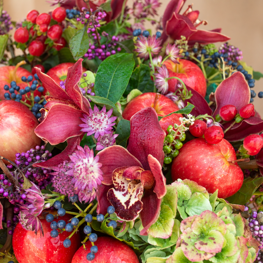 Honeycrisp Harvest arrangement of burgundy Cymbidium orchids, hydrangeas, apples and grapes in a moss-wrapped glass vase