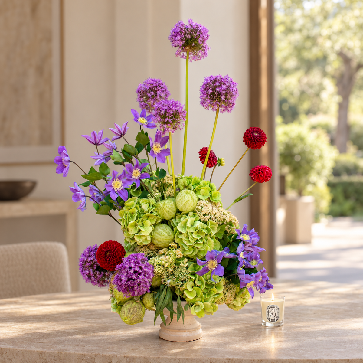 A vibrant floral arrangement in a small cream-colored ceramic pot, featuring purple alliums, green hydrangeas, red globes, and purple clematis, displayed on a stone table in a sunlit patio setting.