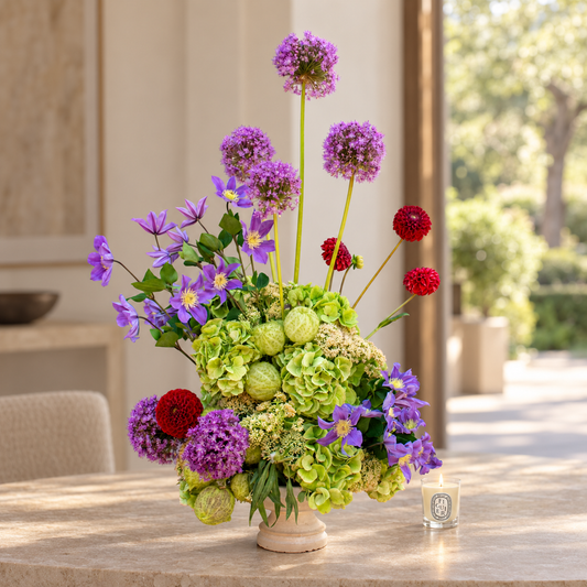 A vibrant floral arrangement in a small cream-colored ceramic pot, featuring purple alliums, green hydrangeas, red globes, and purple clematis, displayed on a stone table in a sunlit patio setting.