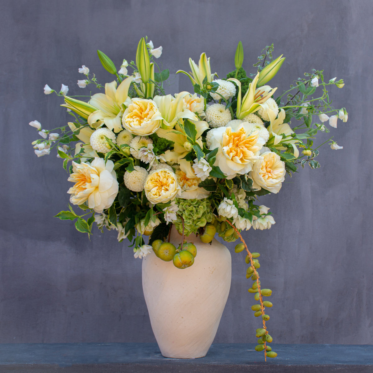 A large floral arrangement featuring pale yellow lilies, cream roses, and green foliage in a textured off-white ceramic vase, set against a dark gray background.